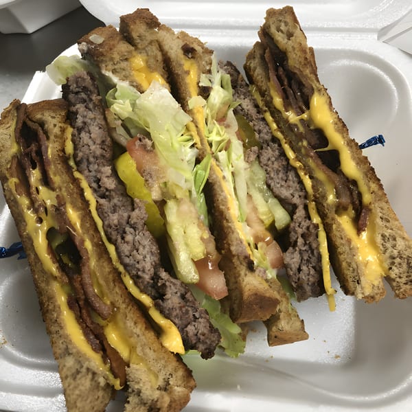 A large Perry Street Grill club sandwich with beef patties, cheese, lettuce, and tomatoes in a styrofoam container.