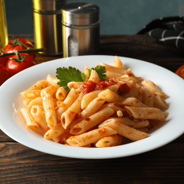 Penne pasta with red sauce and parsley on a white plate, with tomatoes and shakers in the background.