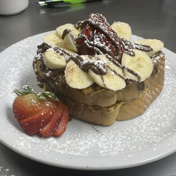French toast stack topped with sliced bananas, chocolate drizzle, powdered sugar, and a side of strawberries.