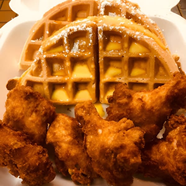 Golden waffles with syrup and powdered sugar, alongside crispy fried chicken pieces in a white container.