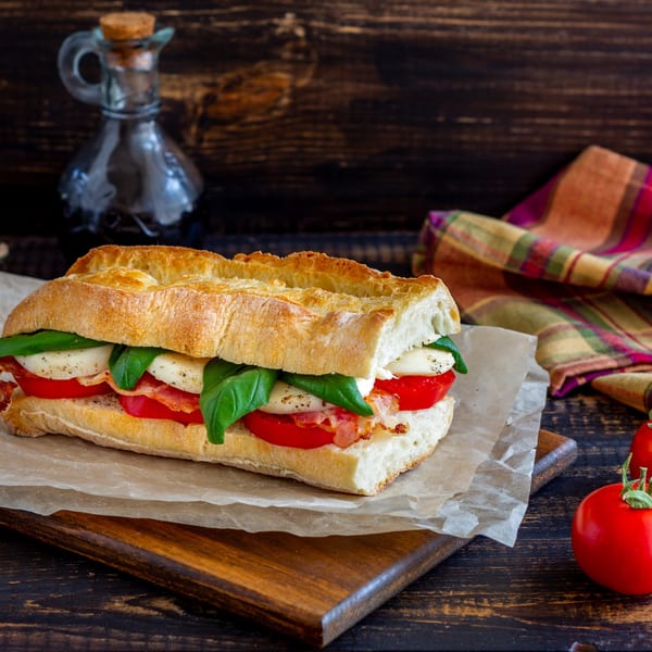 A Caprese sandwich with mozzarella, basil, tomatoes, and bacon on a baguette, with a cruet and tomatoes.